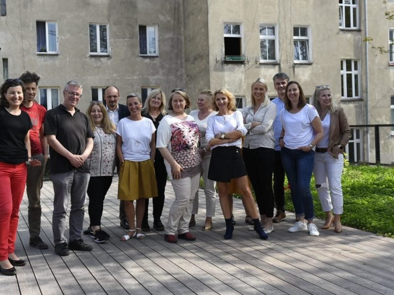 grupa kilkunastu uśmiechniętych osób w różnym wieku i różnych płci (ale więcej kobiet)) stoi na tarasie. W tle okna starych kamienic, po prawej stronie, na dole trawa i niska zieleń.